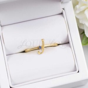 A gold ring with the initial "J" and a blue stone displayed in a jewelry box.