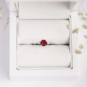 Minimalist silver ring with vibrant round ruby and pointed white side stones, resting in a display case.