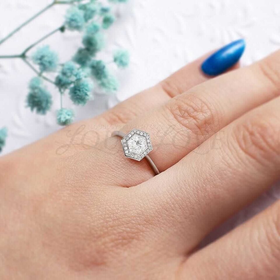 Bezel-set hexagon moissanite with halo of stones in white gold, styled on hand.